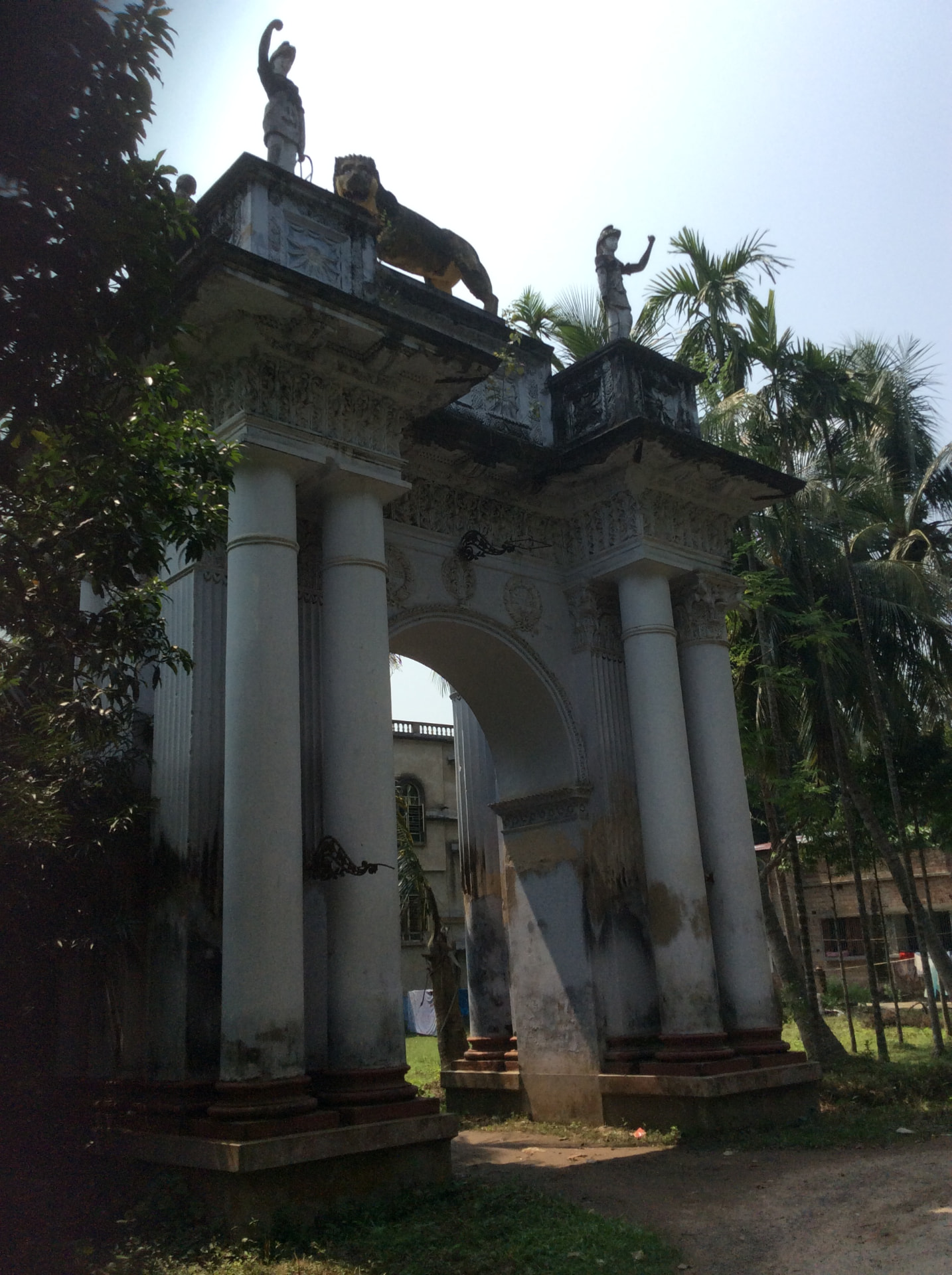 A tall white arched gateway with paired columns, a lion and two standing statues on top, among palm trees, with a large old house seen through the arch