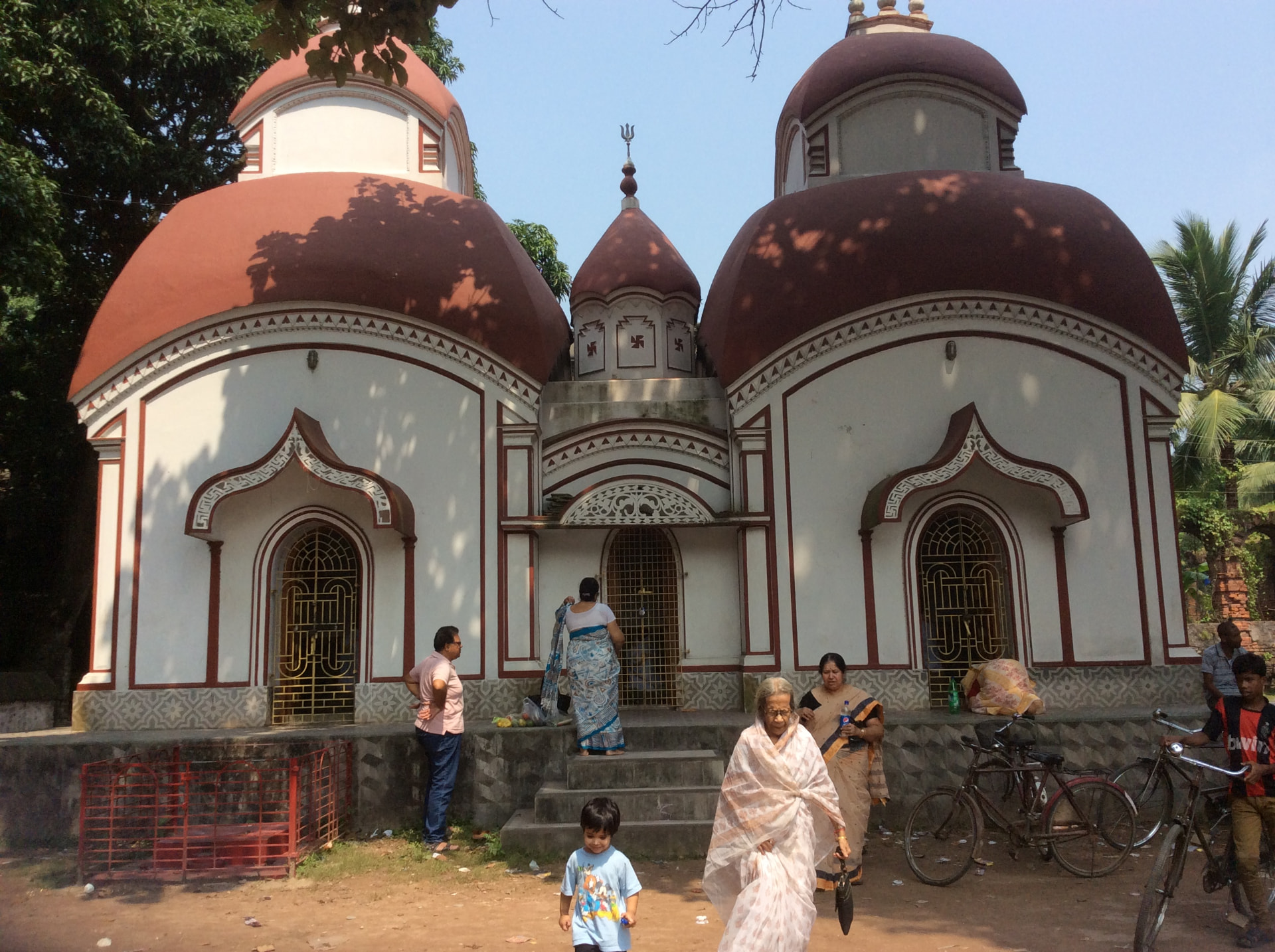 A white temple with two large red domes and a small spire with a trident between them, family members on the steps and in front